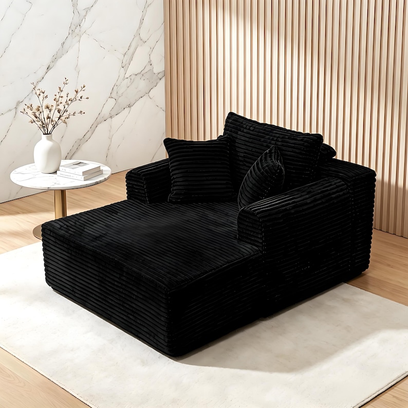 May include: A black corduroy chaise lounge with two matching throw pillows. The lounge has a low profile and a wide armrest. The fabric has a ribbed texture. The chaise is on a white rug.