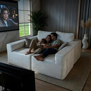 May include: A white corduroy sectional sofa with two people sitting on it. The sofa has a modern design with a ribbed texture. The couple is embracing while watching a movie on a projector screen. A neutral-toned rug and decorative vase are also visible.