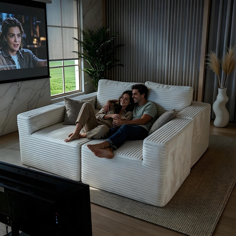 May include: A white corduroy sectional sofa with two people sitting on it. The sofa has a modern design with a ribbed texture. The couple is embracing while watching a movie on a projector screen. A neutral-toned rug and decorative vase are also visible.