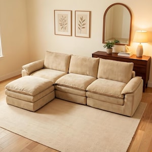 May include: A beige sectional sofa with an attached ottoman sits on a light beige rug. The sofa has large, plush cushions and armrests. Two framed botanical prints hang on the wall, and a wooden cabinet with a mirror and lamp are in the background.