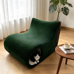 May include: A dark green, plush cat bed with a curved, modern design. The bed has a built-in cave where a black and white cat is resting. A red accent line runs along the side. The cat bed is in a well-lit room with a wooden floor.