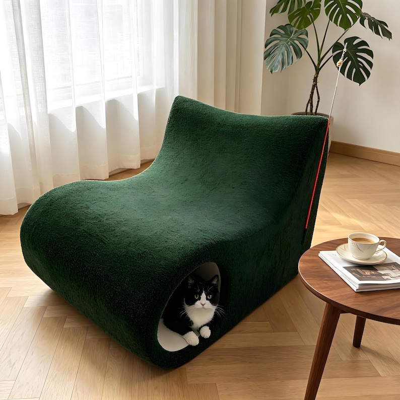 May include: A dark green, plush cat bed with a curved, modern design. The bed has a built-in cave where a black and white cat is resting. A red accent line runs along the side. The cat bed is in a well-lit room with a wooden floor.
