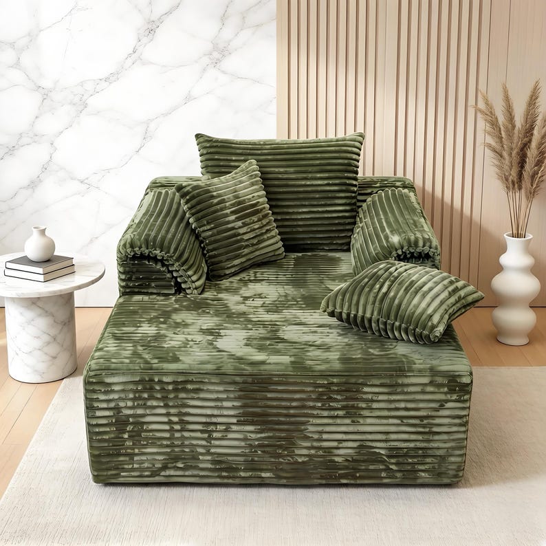 May include: A green corduroy sofa with multiple matching pillows. The sofa and pillows have a ribbed texture. The room has a marble side table with a white vase and a tall white vase with dried plants.