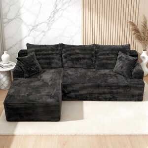 Op de afbeelding: Een donkergrijze hoekbank met een ribfluwelen textuur. De bank heeft een L-vorm met een chaise longue aan de linkerkant en meerdere rugkussens. Twee bijpassende sierkussens zijn zichtbaar. De bank staat op een wit tapijt.