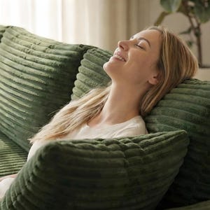 May include: A person is resting on a green corduroy sofa, leaning against matching pillows. The pillows and sofa have a textured, ribbed design. The person is wearing a white shirt and has blonde hair.