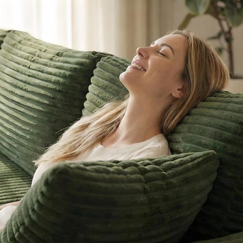 May include: A person is resting on a green corduroy sofa, leaning against matching pillows. The pillows and sofa have a textured, ribbed design. The person is wearing a white shirt and has blonde hair.