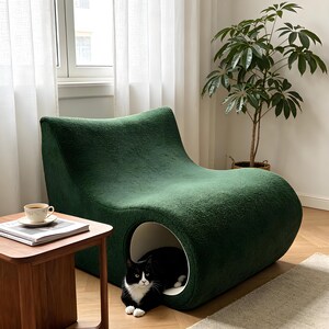 May include: A dark green, modern cat bed with a curved design. The bed has a circular opening on the side, revealing a black and white cat inside. A small wooden side table with a cup and saucer sits nearby.