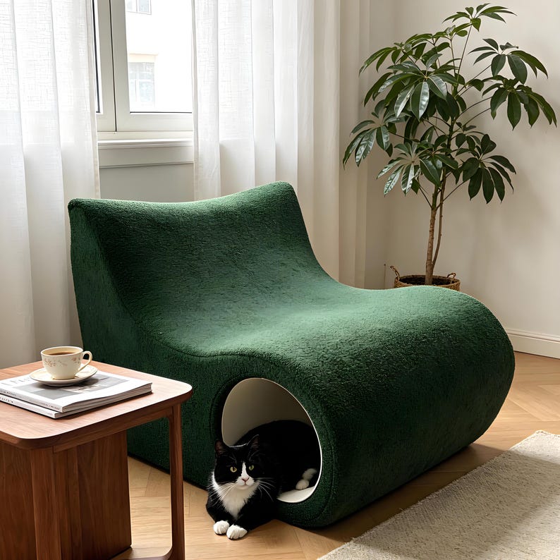 May include: A dark green, modern cat bed with a curved design. The bed has a circular opening on the side, revealing a black and white cat inside. A small wooden side table with a cup and saucer sits nearby.