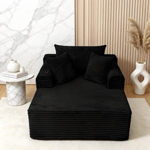 May include: A black corduroy chaise lounge with matching pillows. The lounge has a low profile and is set on a white rug. A marble side table with a white vase and books is to the left.