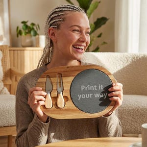 Op de afbeelding: Houten kaasplank met twee kaasmesjes en een leistenen serveergedeelte. Op de plank staat de tekst "Print it your way" op de leisteen. De messen hebben houten handvatten.