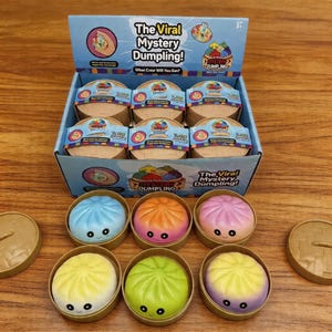 Puede incluir: Una caja expositora de juguetes "The Viral Mystery Dumpling!", con figuras en forma de dumpling sonrientes y coloridas. La caja incluye contenedores individuales de dumpling y el texto "What Color Will You Get?" en la parte delantera. Los dumplings son de varios colores.