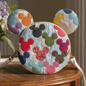 May include: A round, light blue quilted crossbody bag with Mickey Mouse head shapes in various colours. The bag has two rounded ear-shaped accents on top. The bag has a cream-coloured strap and gold-coloured hardware. The bag is sitting on a wooden surface.