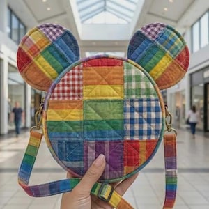 May include: A vibrant, circular crossbody bag with Mickey Mouse ear-shaped details. The bag showcases a patchwork design in rainbow hues, including red, orange, yellow, green, blue, and purple. The strap and ears also feature a rainbow pattern.