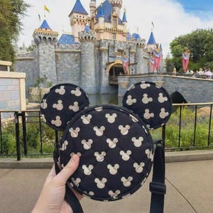K&ouml;nnte beinhalten: Eine schwarze, runde Umh&auml;ngetasche mit Mickey Mouse-Kopfmuster und passenden Ohren. Die Tasche hat einen schwarzen Riemen und wird vor einem Schloss gehalten.