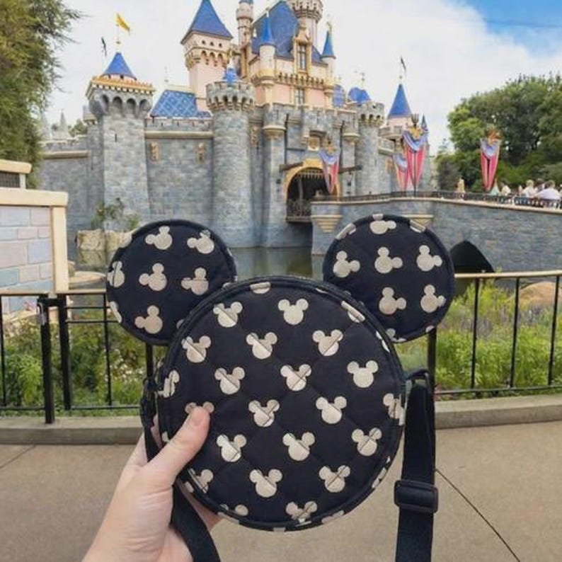 K&ouml;nnte beinhalten: Eine schwarze, runde Umh&auml;ngetasche mit Mickey Mouse-Kopfmuster und passenden Ohren. Die Tasche hat einen schwarzen Riemen und wird vor einem Schloss gehalten.