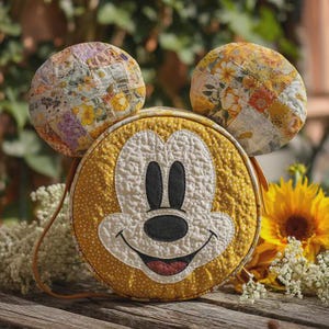 May include: A yellow quilted crossbody bag featuring Mickey Mouse's face, with round ears on top. The bag has a brown strap and is decorated with a white and yellow floral pattern. The background includes sunflowers and greenery.