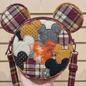 May include: A round, quilted crossbody bag with Mickey Mouse applique details. The bag features a plaid pattern in burgundy, gold, and cream, with coordinating ear-shaped accents. The bag has a zip closure and a burgundy strap.