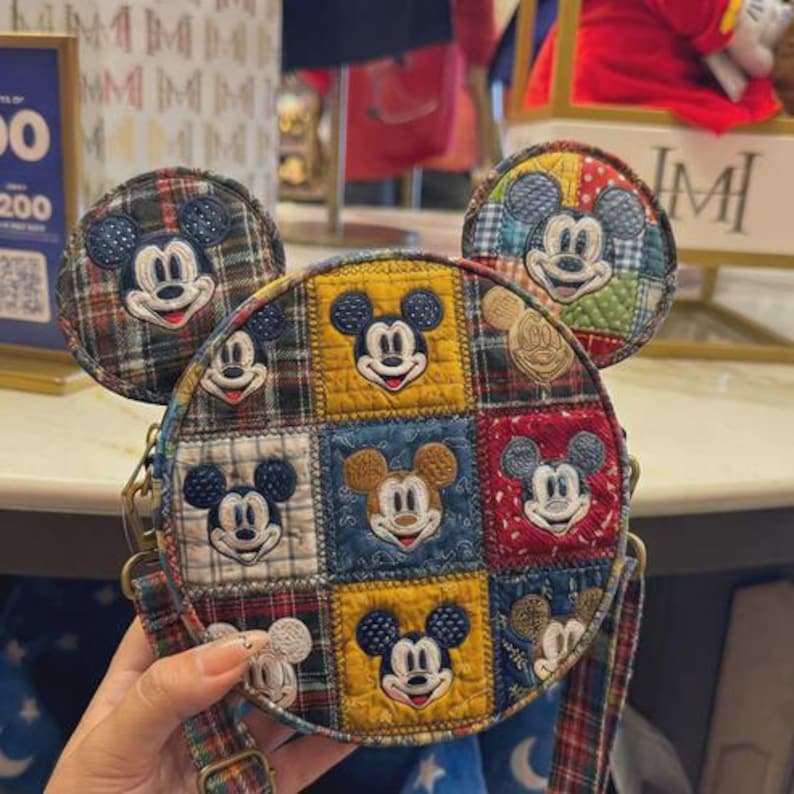 Puede incluir: Un bolso bandolera redondo, estilo patchwork, con Mickey Mouse. El bolso tiene un patr&oacute;n de cuadros con caras de Mickey Mouse bordadas en cada cuadrado. Tiene una correa a juego y dos adornos en forma de orejas de Mickey Mouse.