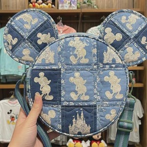 May include: A blue denim-style crossbody bag featuring Mickey Mouse and castle designs. The bag has round ears and a green adjustable strap. The bag is held by a person.
