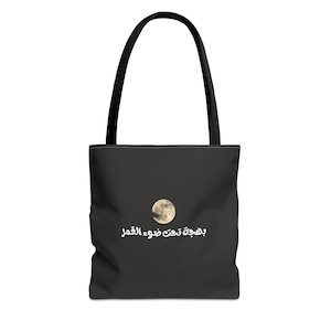 Puede incluir: Bolso tote negro con asas negras. El bolso presenta un gráfico de luna llena y texto árabe. La luna es de color amarillo pálido. El texto es blanco. El bolso está hecho de un material resistente.