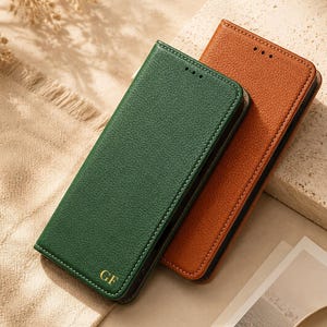 May include: Two rectangular wallet-style phone cases with a textured finish. One case is dark green with the gold embossed letters GF at the bottom, and the other case is tan. Both cases feature a slim profile with visible stitching along the edges.