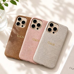 Könnte beinhalten: Drei strukturierte Handyhüllen in Braun, Rosa und Off-White, jeweils mit einem goldenen Monogramm "G&H". Die Hüllen haben einen Kameraausschnitt und sind auf einer weißen Oberfläche angeordnet. Im Hintergrund befindet sich eine Glasvase mit grünen Blättern.
