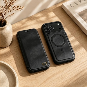 May include: Two black leather-textured phone cases. One case is a folio style with the gold embossed initials GF on the front. The other case features a circular magnetic charging ring on the back and a camera cutout for three lenses.