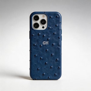 Könnte beinhalten: Eine tiefblaue Smartphone-Hülle mit einer strukturierten Straußenleder-Optik und erhabenen Noppen. In der Mitte ist die Hülle mit den silberfarbenen Großbuchstaben GH personalisiert. Sie verfügt über einen Ausschnitt für ein Dreifach-Kamerasystem.