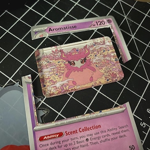Pu&ograve; includere: Un pezzo rettangolare in acrilico con un'illustrazione del personaggio rosa Aromatisse su sfondo floreale, posizionato tra le sezioni superiore e inferiore di una carta collezionabile Aromatisse viola con testo e statistiche visibili.
