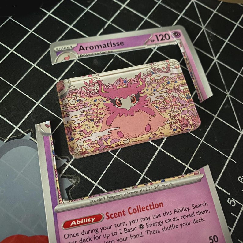 Pu&ograve; includere: Un pezzo rettangolare in acrilico con un'illustrazione del personaggio rosa Aromatisse su sfondo floreale, posizionato tra le sezioni superiore e inferiore di una carta collezionabile Aromatisse viola con testo e statistiche visibili.