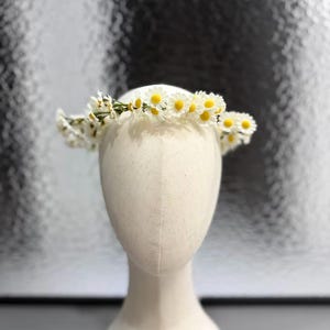 Può includere: Una corona floreale di margherite bianche con centri gialli, esposta su una testa di manichino. La corona floreale è composta da fiori piccoli e delicati e steli verdi. Lo sfondo è una superficie argentata strutturata.