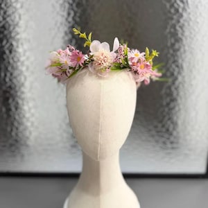 Puede incluir: Una corona floral con flores rosas y blancas y detalles verdes, exhibida en una cabeza de maniquí. Las flores están dispuestas en un patrón circular, creando un accesorio delicado y femenino, ideal para bodas.