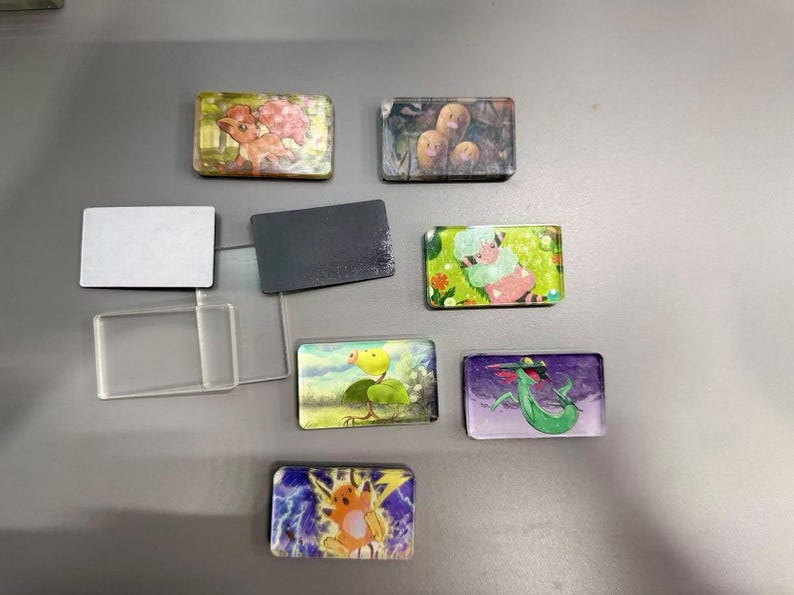 Pu&ograve; includere: Una collezione di magneti rettangolari con varie illustrazioni colorate di personaggi dei cartoni animati, tra cui una creatura simile a una volpe, tre figure marroni, un essere vegetale verde e una creatura elettrica gialla, accanto a cornici in plastica trasparente.