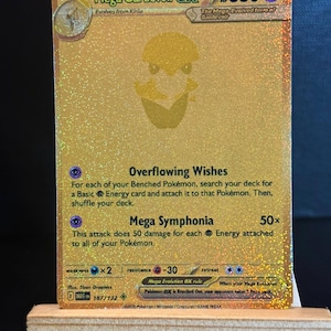 Könnte beinhalten: Eine goldfarbene Mega Gardevoir EX Pokémon-Sammelkarte. Die Karte hat ein holografisches Design mit dem Text "Mega Gardevoir EX" und "HP 360". Die Karte enthält auch Spielanweisungen und den Text "Overflowing Wishes" und "Mega Symphonia".