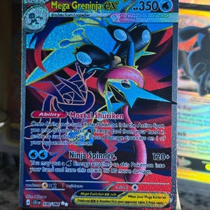 Könnte beinhalten: Eine holografische Mega Greninja EX Sammelkarte mit 350 KP, die eine dunkelblaue und rote Illustration der Figur zeigt. Die Karte enthält den Text zur Fähigkeit Mortal Shuriken und zum Angriff Ninja Spinner, Set-Nummer 100/086.