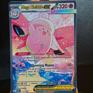 May include: A Mega Clefable EX Pokémon card. The card features a pink Clefable with a white and pink design, and the text "Mega Clefable EX" at the top. The card has the text "Luminous Wing" and "Shooting Moons" with a 120+ damage indicator.