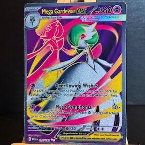 Könnte beinhalten: Eine Pokémon-Sammelkarte von Mega Gardevoir EX. Die Karte zeigt eine holografische Illustration von Gardevoir in Pink-, Lila- und Grüntönen. Text enthält "Overflowing Wishes" und "Mega Symphonia". Die Karte wird auf einer kleinen Holzstaffelei präsentiert.