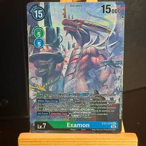 Op de afbeelding: Een holografische Digimon-verzamelkaart met Examon, een draakachtig wezen met een rood en zilver kleurenschema. De kaart toont een speelkost van 15 en een DP van 15.000. De kaart is een Mega-kaart van level 7.