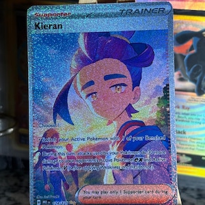 May include: A holographic Kieran Supporter Trainer trading card featuring a stylised illustration of a character with dark purple hair against a sunset background. The card text includes game instructions and the card number 174/131.