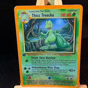 Op de afbeelding: Een Thicc Treecko Pokémon-kaart met een groene en oranje rand. De kaart toont een groen, hagedisachtig wezen met de tekst "Thicc Treecko" en "HP 40". De kaart wordt gepresenteerd op een klein houten ezel.