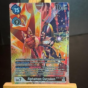 Peut inclure: Carte de jeu Digimon avec Alphamon: Ouryuken. Le personnage central porte une armure noire et dorée sur un fond rouge et orange. On peut lire "Digimon" et "16,000 DP". La carte est présentée sur un chevalet en bois.