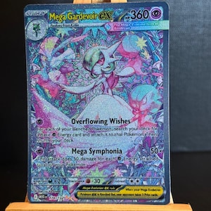 Könnte beinhalten: Eine holografische Mega Gardevoir EX Pokémon-Sammelkarte. Die Karte zeigt eine detaillierte Illustration von Gardevoir im Buntglasstil mit rosa, blauen und weißen Farben. Die Karte wird auf einer kleinen Holzstaffelei präsentiert.