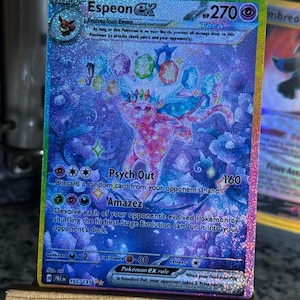 May include: A holographic Espeon ex trading card with a 270 HP rating. The card features a pink Espeon character surrounded by colorful floating gems and blue floral patterns. Text includes Psych Out and Amazez attacks, with the card number 155/131.