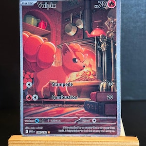 Könnte beinhalten: Eine Vulpix Pokémon-Sammelkarte. Die Karte zeigt ein rotes fuchsähnliches Wesen vor einem Kamin. Die Karte trägt die Aufschriften "Vulpix", "Stampede" und "Combustion". Die Karte wird auf einer kleinen Holzstaffelei präsentiert.