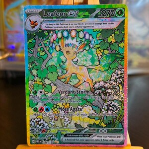 May include: A Leafeon ex Tera trading card featuring a holographic finish, 270 HP, and artwork of the creature surrounded by colourful gems and lush foliage. The card displays the attacks Verdant Storm and Moss Agate with detailed text.