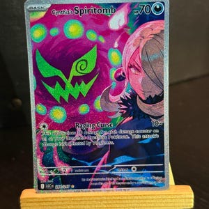 以下が含まれることがあります： 「Cynthia's Spiritomb」をフィーチャーしたホログラフィックトレーディングカード。カードは、キャラクターイラストが描かれた紫と緑の背景です。カードには「Raging Curse」と「700 HP」の文字も記載されています。