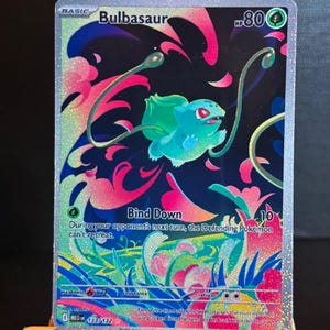 Könnte beinhalten: Holografische Bulbasaur Pokémon-Sammelkarte. Die Karte zeigt ein grünes Bulbasaur mit rosa und blauen floralen Akzenten vor einem dunklen Hintergrund. Text enthält "Bulbasaur", "Bind Down" und weitere Spieldetails.