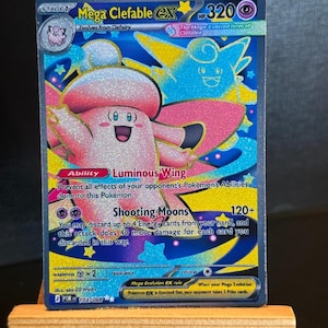 May include: A Mega Clefable EX Pokémon card. The card features a pink and white Clefable with a blue background and star accents. The card has the text "Shooting Moons" and "Luminous Wing".