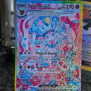 Könnte beinhalten: Eine holografische Sylveon ex Sammelkarte mit einer Tera-Krone, einer rosa und blauen Farbpalette sowie dem Text Magical Charm und Angelite. Die Karte zeigt 270 KP, eine Phase-1-Entwicklung und enthält detaillierte Spielmechaniken.