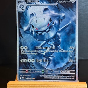 以下が含まれることがあります： ホログラフィックのSteelixポケモンカード。カードには、赤い目のメタリックグレーのSteelixが、青とグレーの背景に描かれています。「Welcoming Tail」や「Skull Bash」などのテキストがカードに表示されています。